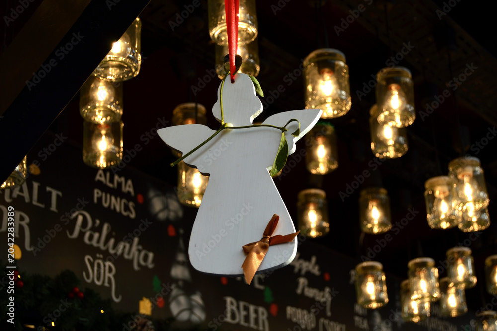 Naklejka premium Christmas market scene, angel ornament hanging, warm festive lights, cozy winter atmosphere, European holiday fair, seasonal street decoration, gift shopping mood, evening bokeh glow.