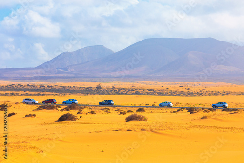 Fotografie Corralejo sand dunes of Fuerteventura provide a stunning desert backdrop for convoy of vehicles driving along a scenic coastal road
