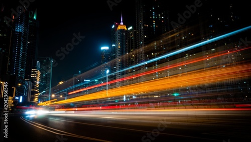Obraz na plátně Dramatic cinematic long-exposure night shot of orange, red and cyan light trails blazing along a city highway, framed by illuminated glass skyscrapers towering against the dark sky
