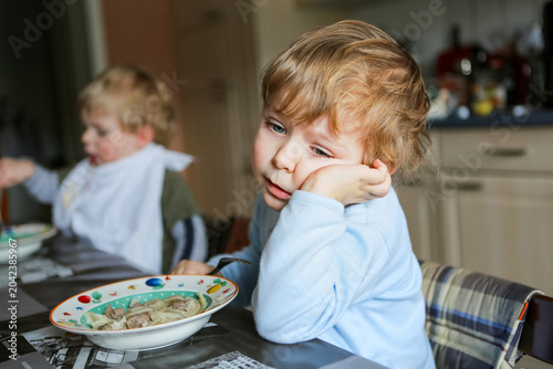 Two little boys eating meal in kitchen at home or daycare nursery, siblings enjoying food together at table, candid family lifestyle moment, real life, real people. Sad child