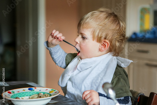 Little boy eating meal in kitchen at home, child enjoying food with fork and bib in candid family lifestyle scene, real life, real people.
