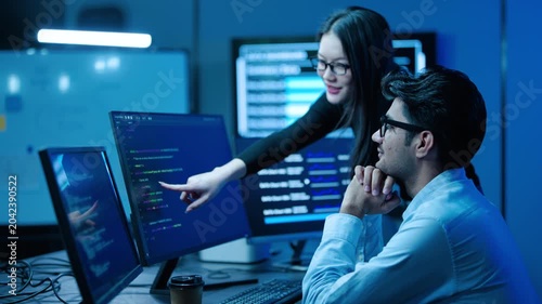 Two software engineers collaborate on computer coding projects in a high-tech office at night. A female programmer points to a screen, discussing application development with her male colleague.
