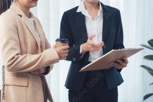 Canvastavla Business Meeting Discussion: Two professional women in business attire engage in a serious discussion, one holding a coffee cup and the other a clipboard, in a well-lit office setting