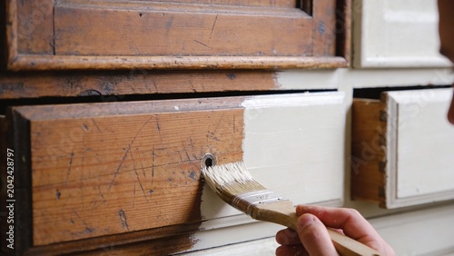 Furniture restoration project showing hand applying white paint with brush onto vintage wooden cabinet surface transforming worn scratched wood into refreshed shabby chic finish DIY home improvement 