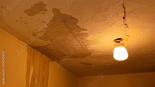 Damaged ceiling with water stains and cracks, illuminated by a single light bulb. A close-up view of a neglected ceiling showing significant water damage, mold growth, and structural cracks.