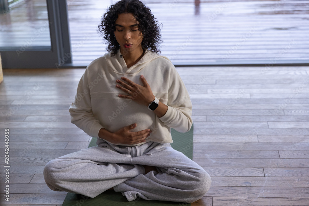 Obraz premium Non-binary person meditating cross-legged on green mat by glass door, wearing cream hoodie
