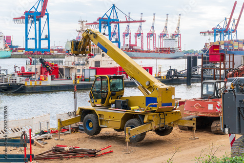 A yellow mobile crane works on a sandy construction site beside a harbor, with cargo ships, tugboats, container stacks, and gantry cranes in the background. Strong image for port logistics, heavy mach