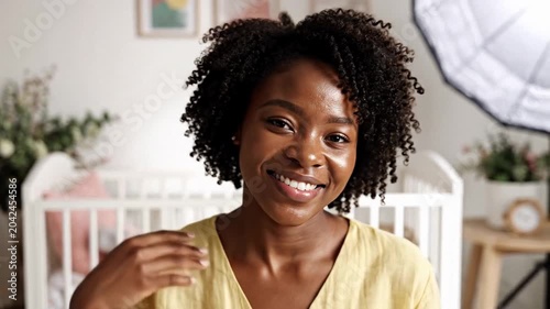 Smiling woman with natural curly hair in a bright nursery, soft daylight and crib behind her, warm welcoming mood for parenting and lifestyle themes