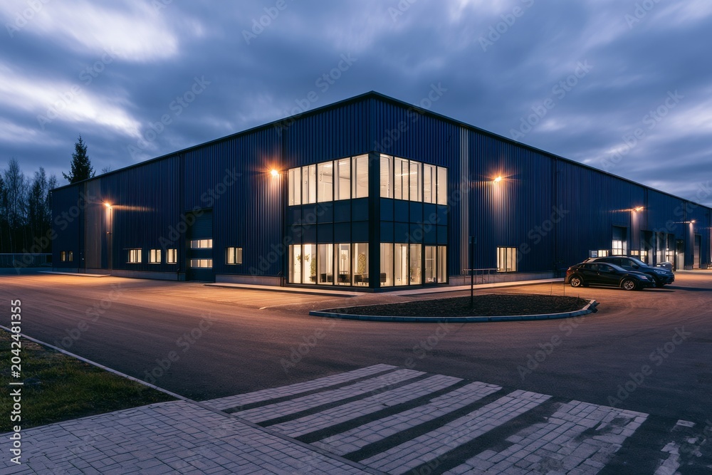 Fototapeta premium Night view of a modern warehouse building with parking area and street lights in an industrial zone