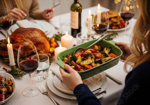 Closeup of a person serving roasted vegetables at a festive holiday dinner table with a roasted turkey, wine, and candles