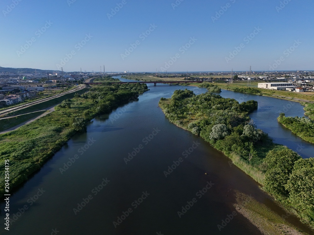 Fototapeta premium Aerial view of calm river winding through green landscape. Blue sky stretches above serene waterway and trees. Urban skyline blends with natural scenery in distance. Ideal for environmental, travel