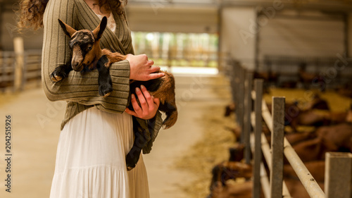 Photos Woman holding a newborn goat in a barn with other goats in the background