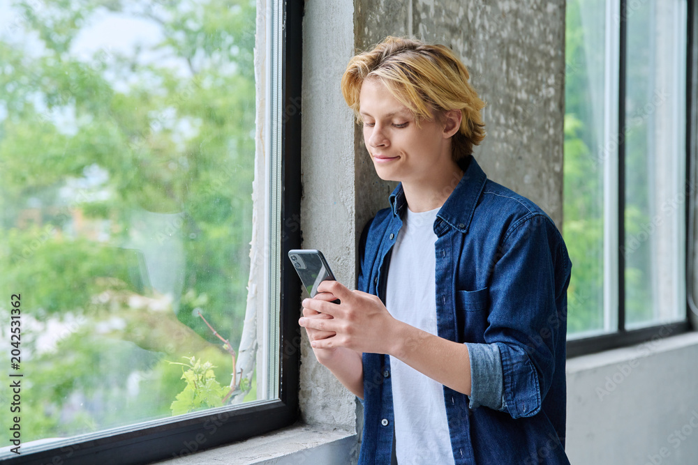 Fototapeta premium Smiling teenage boy using a smartphone by window