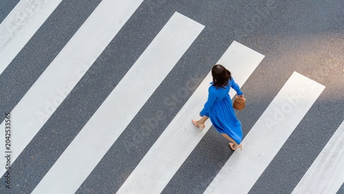 Obraz na plátně Overhead top-down view of a young woman in a vivid blue dress with a wicker bag walking confidently across a wide white zebra crosswalk on a grey urban asphalt surface