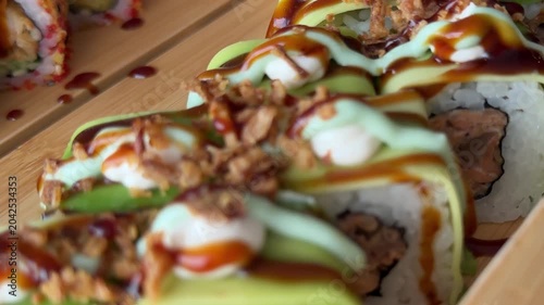 Fresh sushi rolls wrapped with avocado and topped with creamy sauce rest beside bright green wasabi on a decorative wooden serving boat.