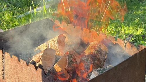 Outdoor barbecue preparation with firewood burning inside metal grill while people ignite campfire and add different wood for cooking atmosphere.