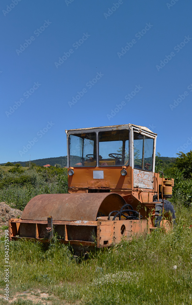 Obraz premium Abandoned vintage road roller in a grassy field with clear blue sky and distant hills visible in the background