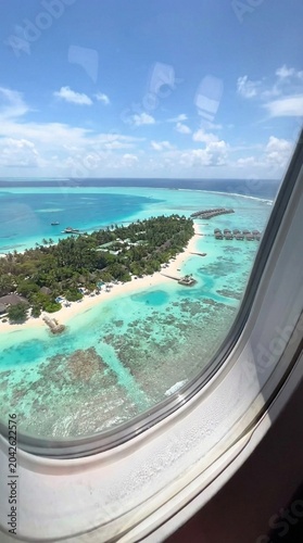 Aerial view through airplane window shows tropical island with white sand beaches and turquoise lagoon, bright sunny sky and calm water, evokes travel escape and vacation mood