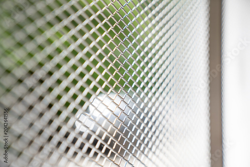 Metal mesh screen with diamond shaped openings. Silver grid pattern looks clean, modern, and strong, with soft green outdoor blur behind it.