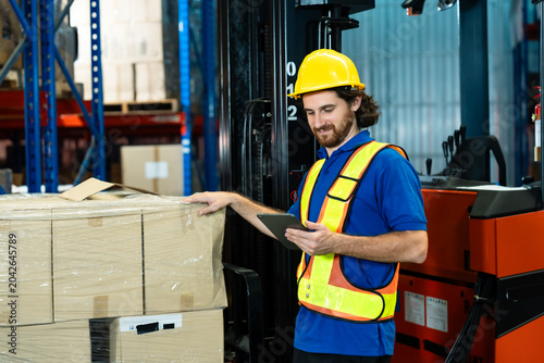 warehouse man in helmet vest using digital tablet to verify cargo quantity with hand placed on pallet of packed boxes near forklift in industrial storage area during routine inspection