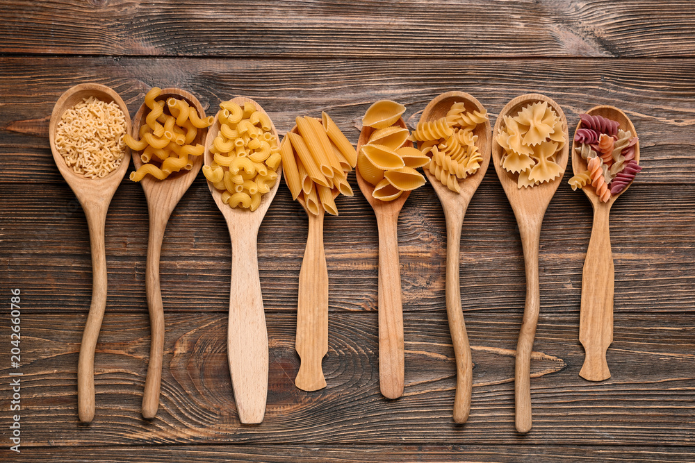 Obraz premium Wooden spoons with different types of pasta on wooden background