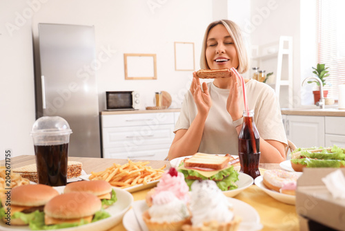 Beautiful woman at table full of unhealthy food in kitchen. Overeating concept