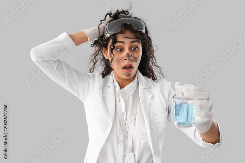 Shocked female African-American chemist with flask on light background