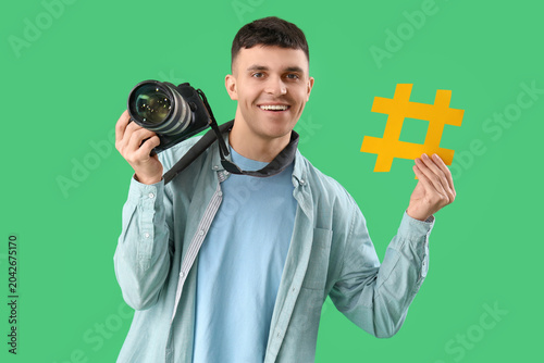 Handsome male photographer with photo camera and hashtag sign on green backgr...