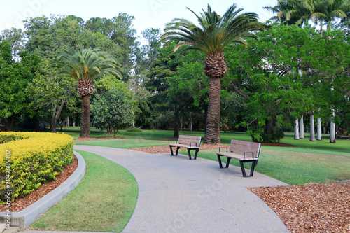 Two wooden park benches alongside a curved concrete footpath surrounded by lush green lawns and palm trees at the Rockhampton Botanic Gardens, Queensland, Australia