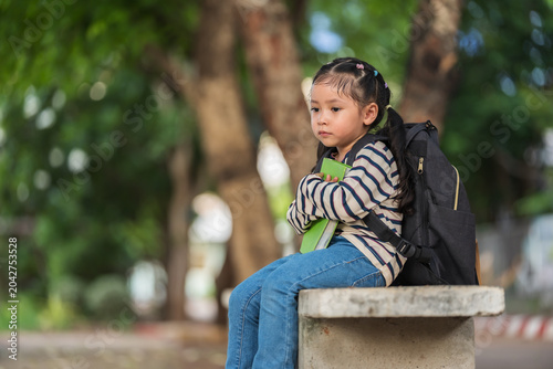 worried child girl sitting on a concrete bench outdoors of school with a book. Education and learning concept.
