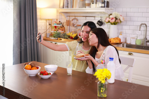 Two young Asian women enjoying healthy breakfast, eating fruit and bread while taking a selfie with a smartphone in a bright kitchen.
