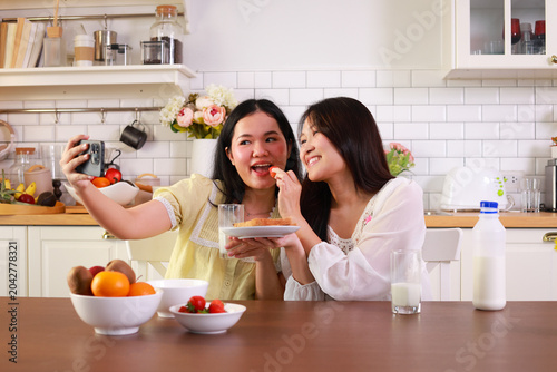 Two young Asian women enjoying healthy breakfast, eating fruit and bread while taking a selfie with a smartphone in a bright kitchen.