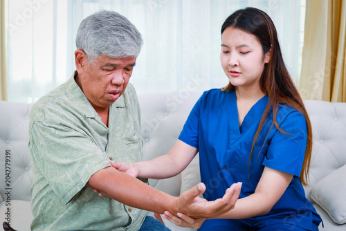 A female physiotherapist treating a senior man with arm and elbow pain on a sofa at home. Professional healthcare support for elderly rehabilitation and physical therapy.