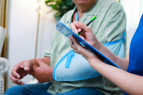 Close up of a female doctor or nurse helping an injured senior man with an arm injury to fill out a health checkup or consent form.