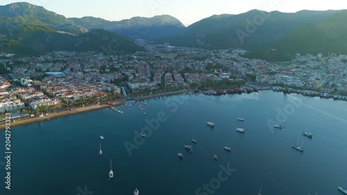 Marmaris, Turkey. Aerial drone view over the bay at sunrise, golden light cutting across pine-covered mountains and terracotta rooftops with yachts anchored in deep blue water. Aerial View