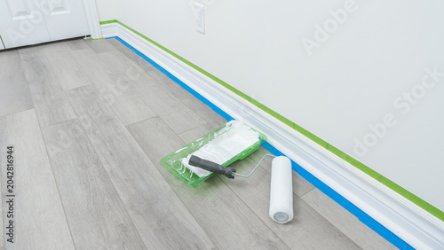 Home renovation scene featuring paint roller, tray, and painter's tape on freshly prepared floor with white walls and door frame visible