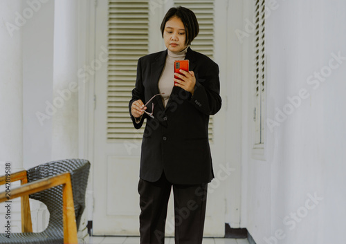Asian Businesswoman short hair in black suit holding a cell phone takes off her glasses standing in room.