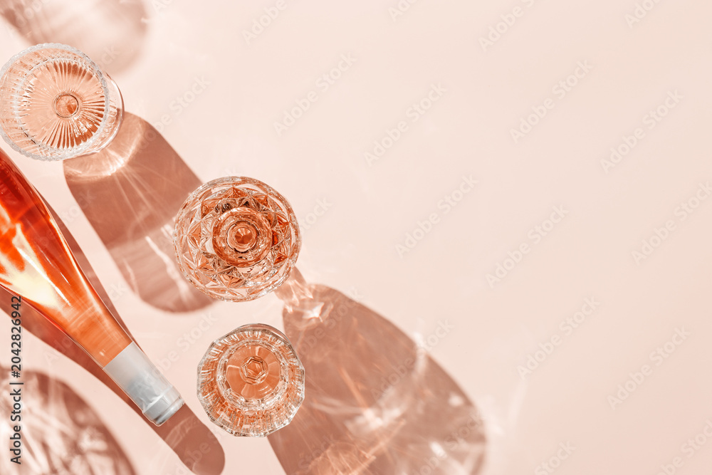 Fototapeta premium Wine Glasses and bottle of rose wine on pink background, sun glare and reflection from sun. Aesthetic composition from crystal glasses, sparkling alcohol drink, champagne, Summer romantic setting