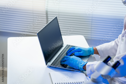 Fotografía Close view of a healthcare worker in blue gloves typing on a laptop at a white desk with a notebook, showing medical research, telehealth, data entry, and clean clinical work