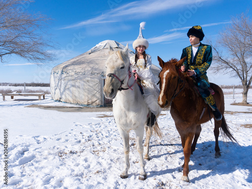 Fotografie Kazakh man and woman in traditional clothing standing with horses near a yurt, representing nomadic culture and heritage of Kazakhstan