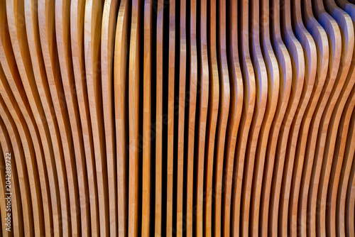 A full-frame shot of a decorative architectural wall feature consisting of numerous vertical, closely arranged wooden slats with unique curved profiles, creating a rhythmic wave-like pattern.