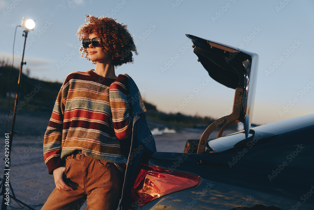 Fototapeta premium Woman lifestyle portrait with curly hair wearing sunglasses beside a car trunk, laptop open with a graduation cap, outdoors at sunset, casual travel mood, soft evening light, natural breeze