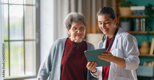 Smiling Female Doctor Consults Happy Senior Woman at Home with Digital Tablet