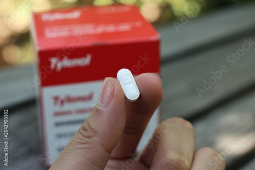Hand holding white caplet medicine pill with a blurred pharmaceutical box in the background