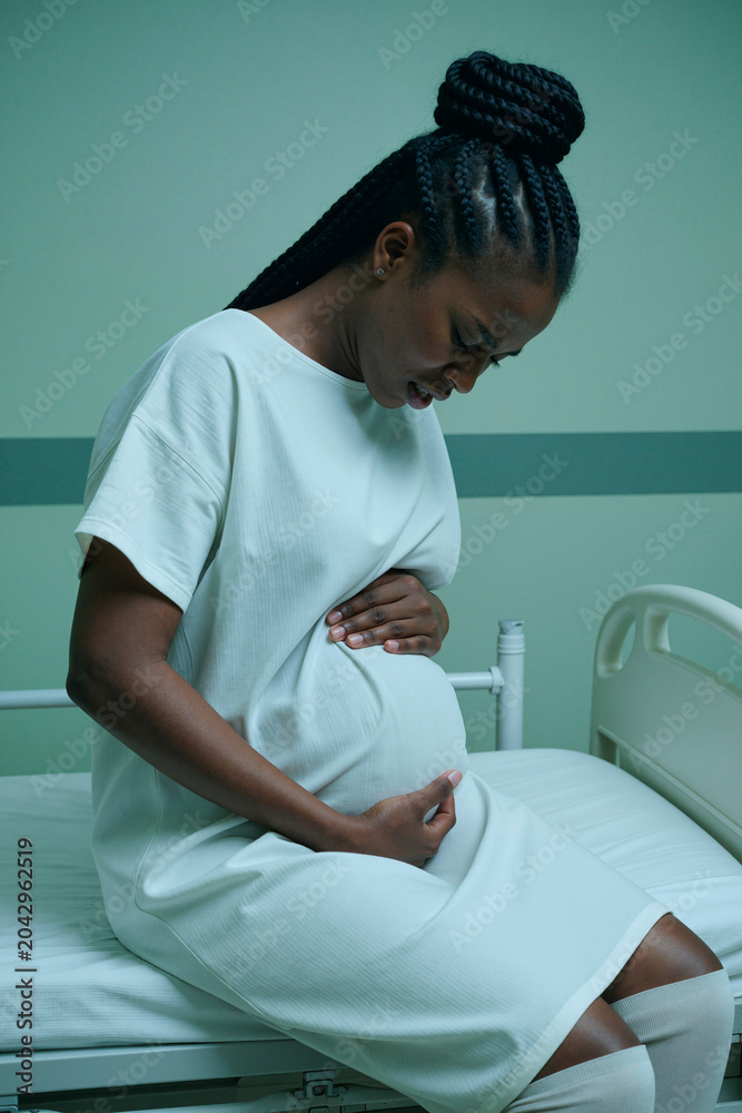 Fototapeta premium Vertical shot of pregnant Black woman sitting on hospital bed, holding belly and feeling discomfort during labor assessment. Ideal for maternity care, prenatal health, obstetrics marketing