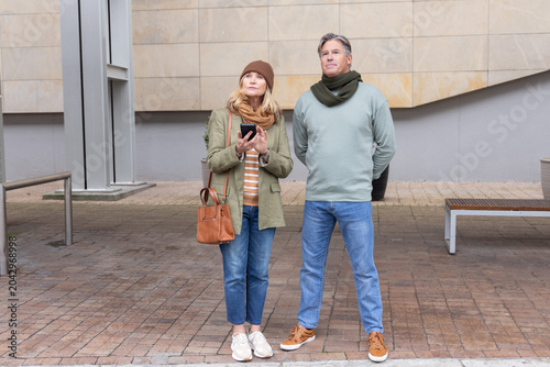 Senior couple standing on paved plaza in front of modern building holding smartphone and handbag
