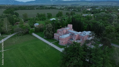 Aerial view of Chateau Mukhrani royal palace and estate in Georgia, magnificent pink castle architecture with a tower surrounded by a lush green park, gardens and pathways on a spring day.