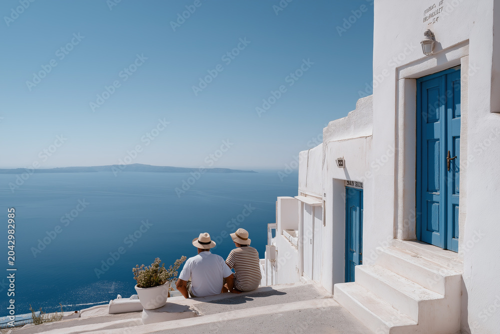 Obraz premium Couple in straw hats sitting beside iconic blue-doored white Santorini building overlooking serene Aegean Sea.