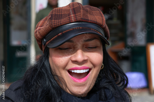 Tableau sur toile A close-up portrait of a laughing woman wearing a brown plaid newsboy cap and vibrant pink lipstick, capturing a cheerful and spontaneous moment in a casual urban environment