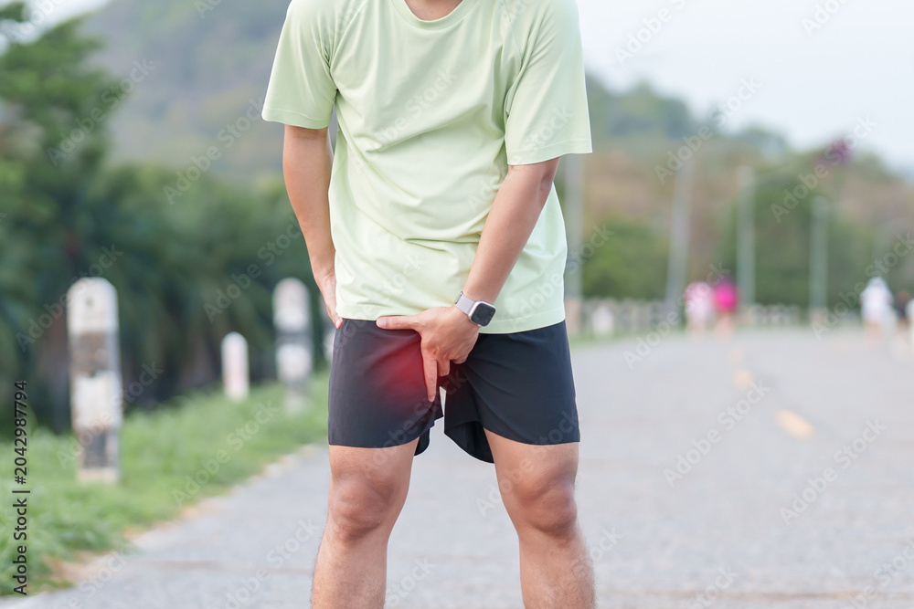 Naklejka premium Young adult male runner suffering from acute groin pull and muscle pain during running workout. Man holding his inner thigh due to sports injury and strain. Medical and healthcare concept